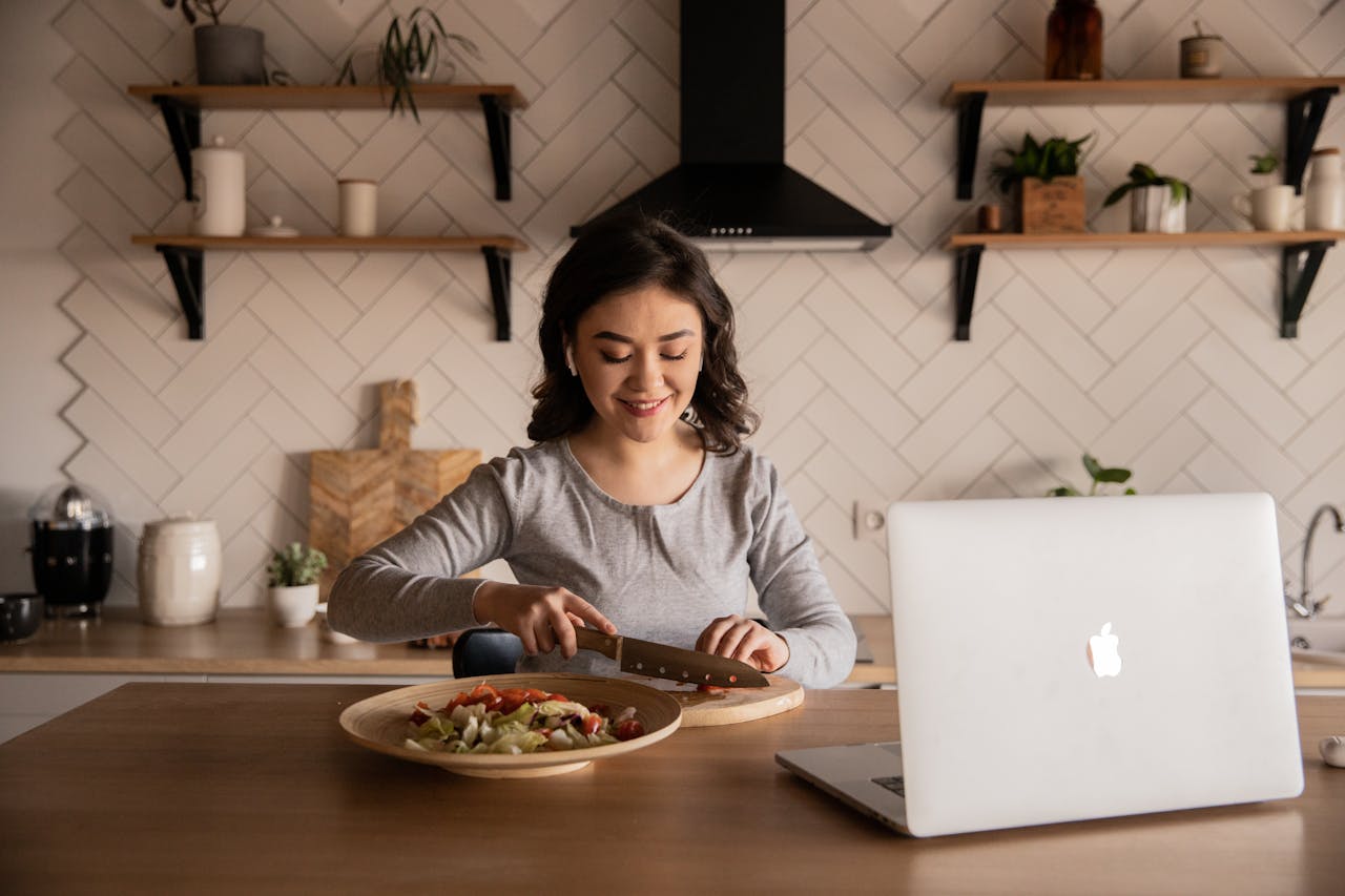 Home Cheerful ethnic female cutting fresh vegetables on cutting board while sitting at wooden table in kitchen with open portable computer in apartment