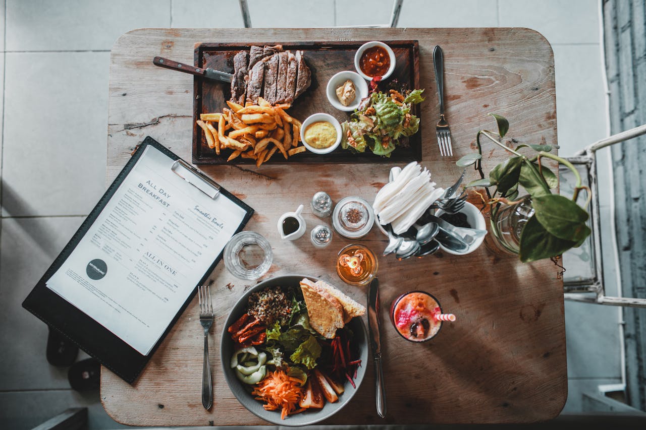 Home Top view of wooden table with salad bowl and fresh drink arranged with tray of appetizing steak and french fries near menu in cozy cafe