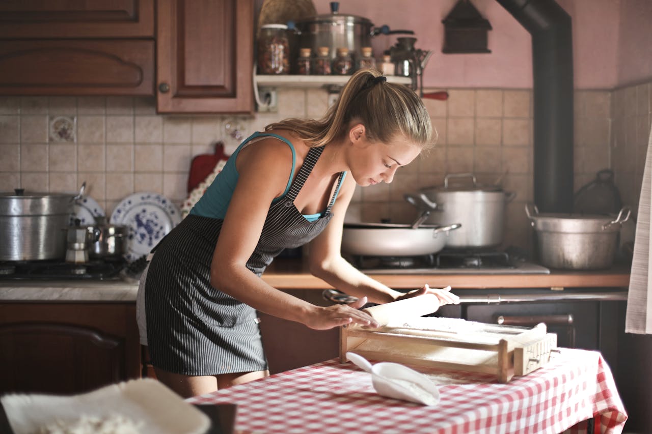 Home Side view of housewife wearing apron standing at table in cozy kitchen and preparing dough for baking while using rolling pin